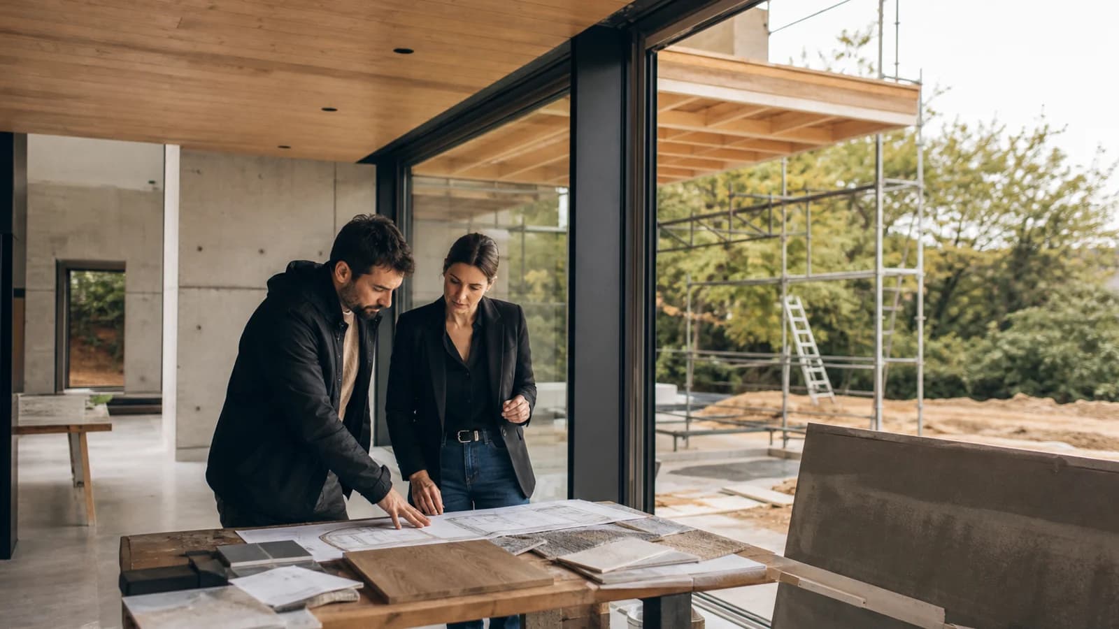 Deux professionnels consultent des plans posés sur une table dans une maison en construction avec échafaudage extérieur.
